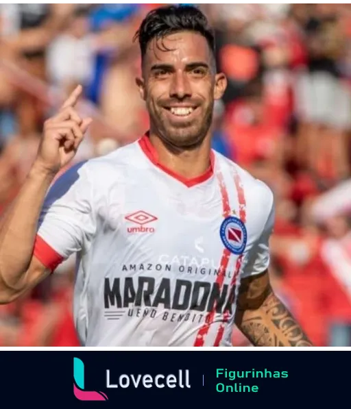 Jogador do Argentinos Juniors sorrindo e comemorando, apontando para cima, vestindo camisa branca com listras vermelhas e logo da Umbro, patrocínio da Amazon Original Maradona visível