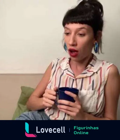 Tina sentada em ambiente caseiro, surpresa, segurando caneca azul, vestindo camisa listrada e com cabelos em coque alto, realçando brincos grandes e coloridos