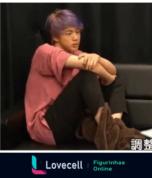 Imagem de um membro do BTS com cabelo roxo e camiseta rosa, sentado abraçando os joelhos em um sofá. No canto inferior direito, há caracteres asiáticos.