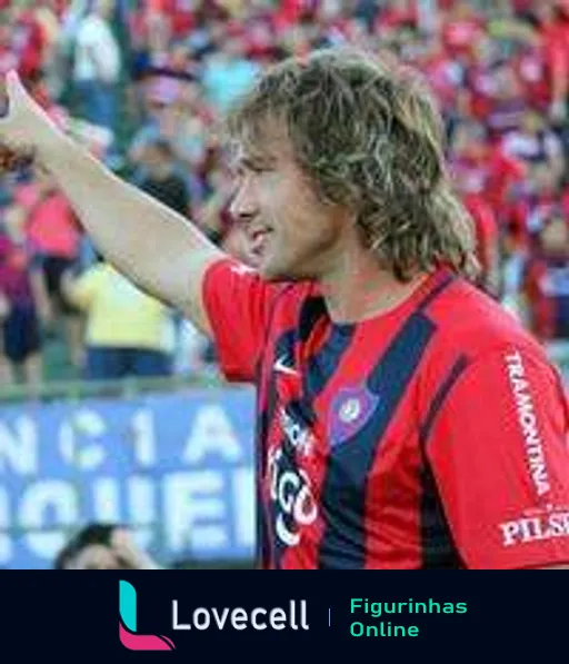 Jogador do Cerro Porteño comemorando gol, levantando a mão direita, com uniforme listrado vermelho e azul, cabelo comprido e desgrenhado, em estádio cheio e vibrante