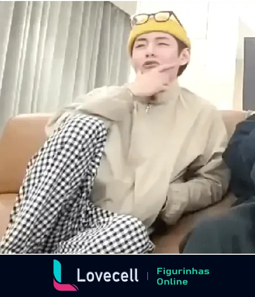 Membro de BTS, sentado de lado no sofá, usando um chapéu amarelo e segurando o queixo pensativamente.