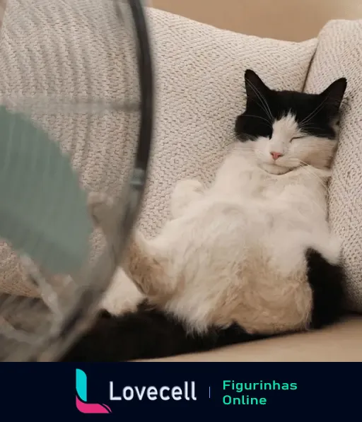 Gato preto e branco sentado descontraidamente no sofá enquanto um ventilador gira ao lado, ajudando a refrescar-se do calor. Pasta: bichinhos com calor.