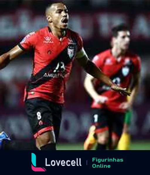 Jogador do Atlético-GO número 8 em uniforme vermelho e preto celebrando com entusiasmo em campo, gritando com estádio ao fundo e um adversário observando