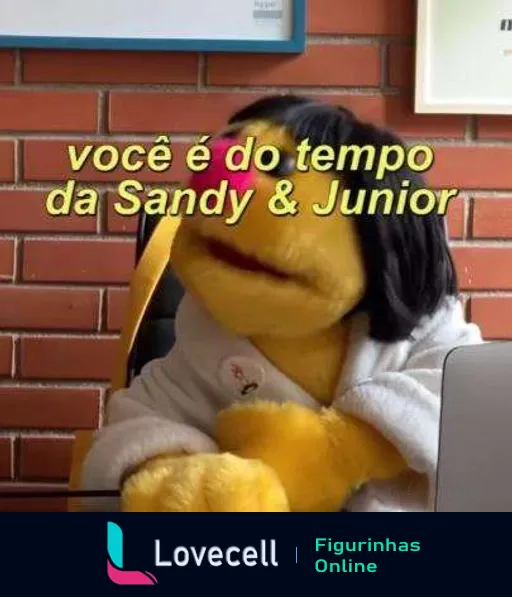 Pelúcia amarela com roupão branco e peruca preta sentada em frente a um laptop, exibindo um ar pensativo e uma mensagem 'você é do tempo da Sandy & Junior'